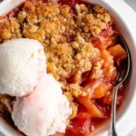 A close-up of golden-brown rhubarb crisp with a buttery oat topping, served warm in a white ceramic dish.