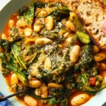 A rustic bowl of Mediterranean white beans and sautéed greens garnished with lemon wedges and fresh herbs.