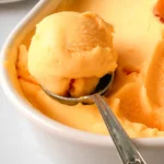 A bowl of homemade peach sorbet, creamy and vibrant, garnished with fresh peach slices and mint leaves, served in a white dish.