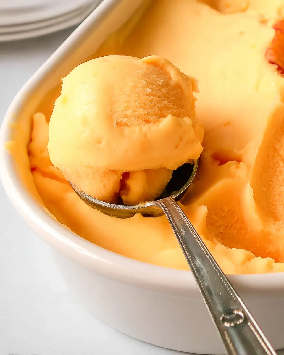 A bowl of homemade peach sorbet, creamy and vibrant, garnished with fresh peach slices and mint leaves, served in a white dish.