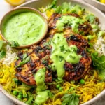 Peruvian-style chicken and rice served with vibrant green sauce, garnished with fresh herbs and lime wedges on a white plate.