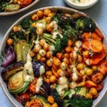 Colorful Roasted Veggie Chickpea Bowls topped with creamy maple tahini dressing, served in a white bowl with quinoa and fresh herbs.