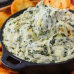 Creamy spinach artichoke dip served warm in a white baking dish, topped with melted golden cheese and ready to be scooped with bread or chips.