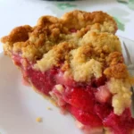 A golden-brown Strawberry Rhubarb Pie with a buttery crumb topping, oozing with juicy fruit filling on a rustic pie plate.