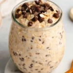 A mason jar filled with creamy cookie dough overnight oats topped with mini chocolate chips and a drizzle of peanut butter, served with a spoon on a rustic wooden surface.