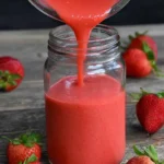 Glass jar of homemade Easy Strawberry Vinaigrette with a vibrant pink hue, surrounded by fresh strawberries and greens.