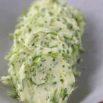 A small dish of creamy garlic scapes butter with flecks of green herbs, next to a bundle of fresh garlic scapes and sliced crusty bread.