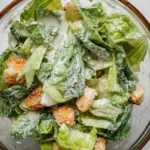 A small glass jar filled with creamy homemade Caesar salad dressing, surrounded by lemon slices, grated Parmesan, garlic cloves, and anchovies on a rustic wooden surface.