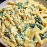 A creamy bowl of spinach artichoke pasta garnished with grated Parmesan, served in a white ceramic bowl on a rustic wooden table.